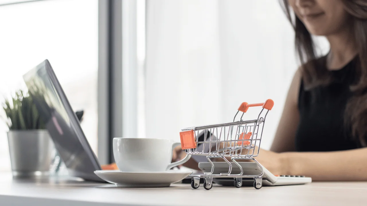 A woman sitting at a desk working on a laptop next to a small miniature shopping cart and coffee cup.
