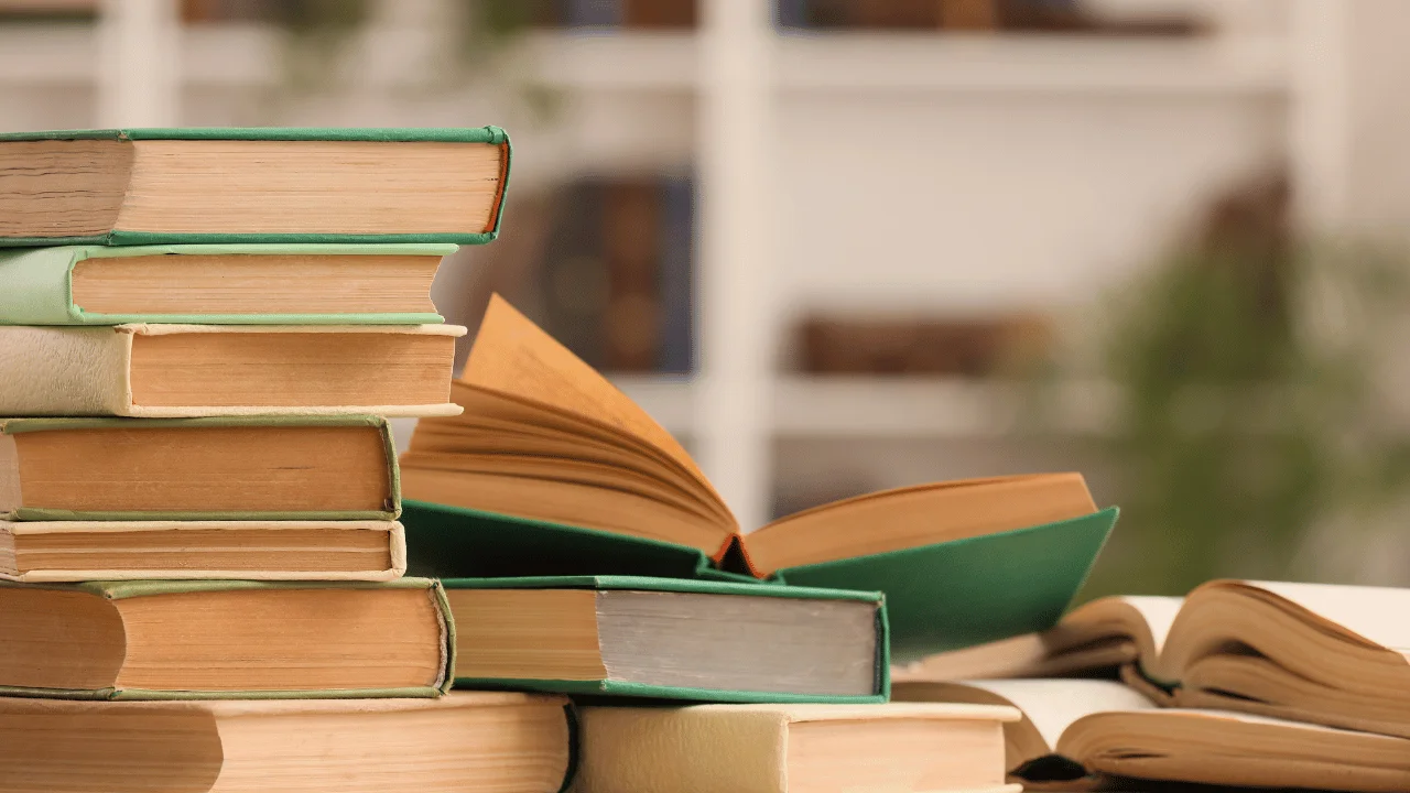 Stacks of books with one open book resting on top against a blurred bookshelf background.