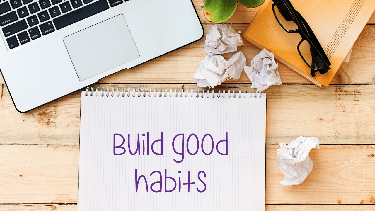 Top-down view of a laptop, glasses, yellow notebook, and a spiral pad with handwritten text reading 'Build good habits'.