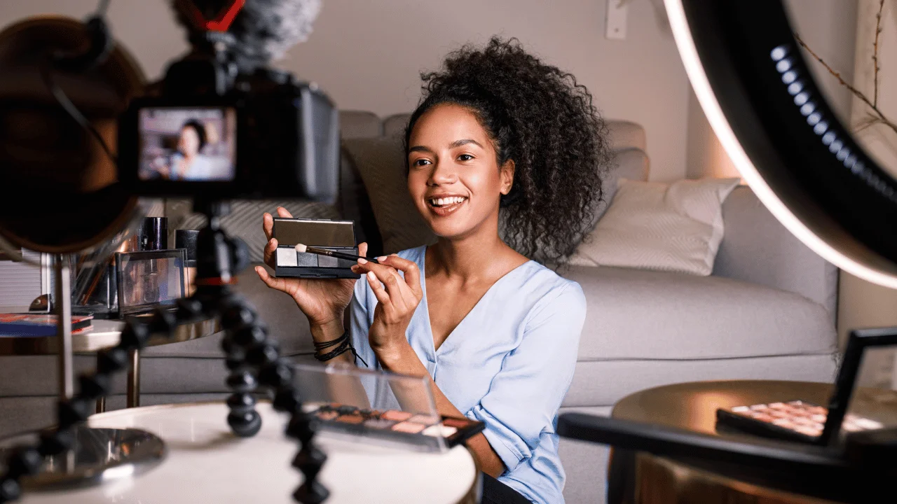 A woman filming a video blog with a camera on a tripod, holding up a small makeup palette in front of a ring light.