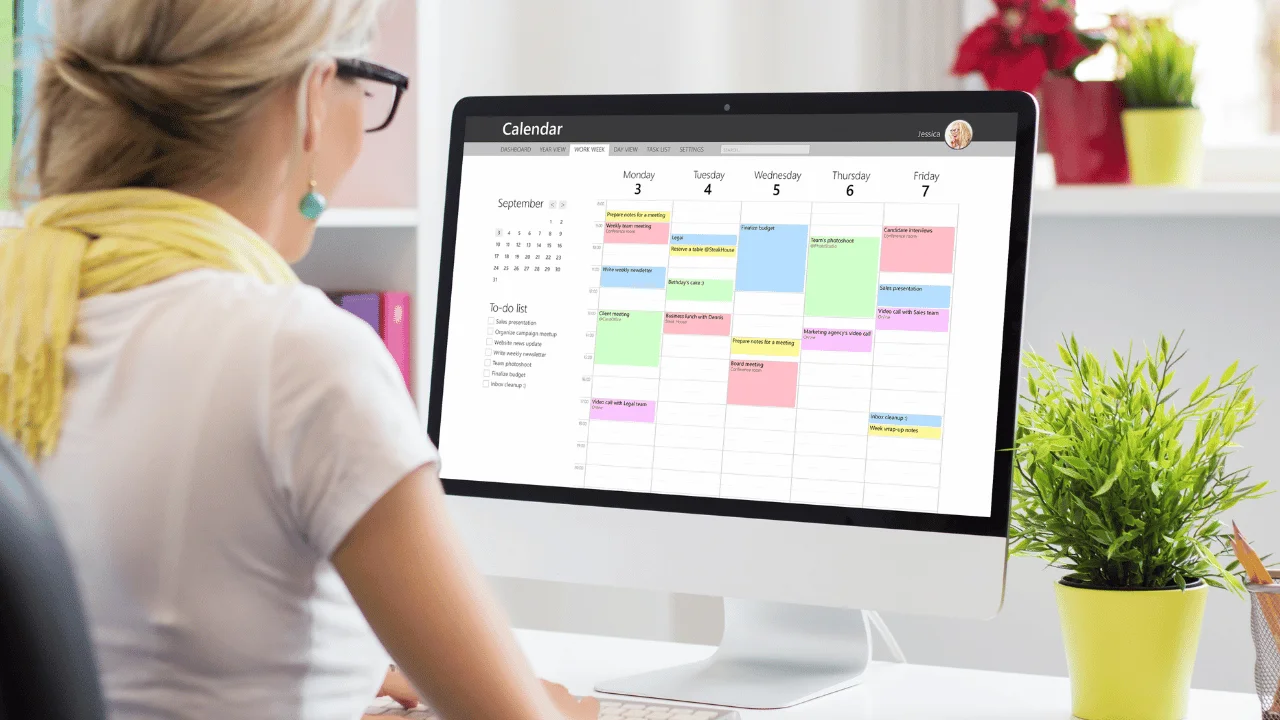 A woman sitting in front of a computer screen displaying a colorful digital calendar schedule.