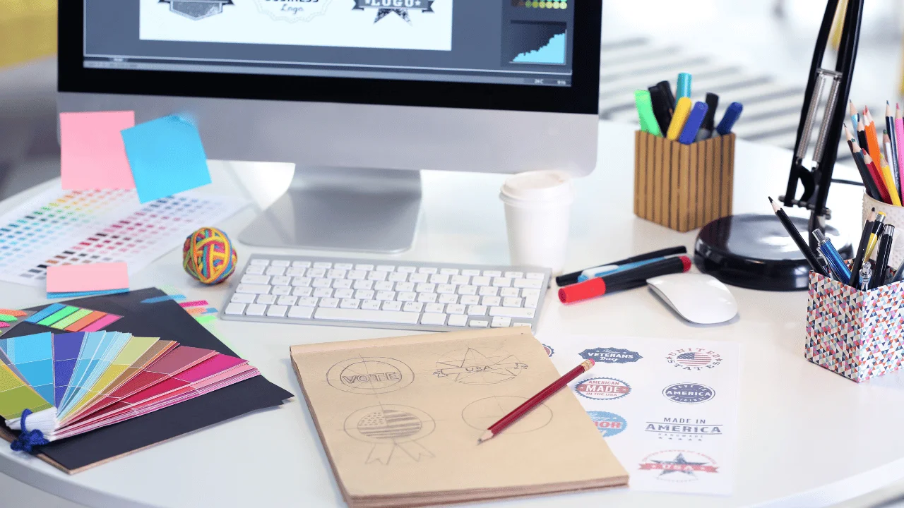 Graphic design workspace featuring a computer screen with design software, color swatches, sticky notes, stationary, and a sketchbook with hand-drawn logo ideas.