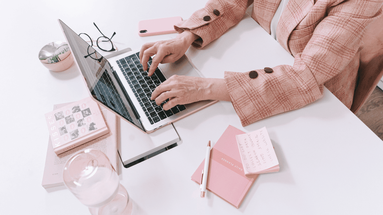 Overhead view of a person typing on a laptop at a bright desk surrounded by pink stationery and notebooks.