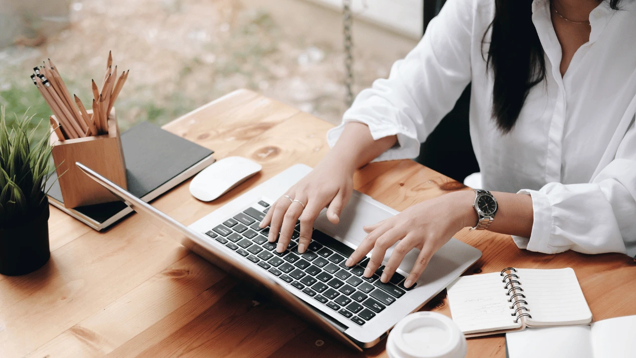 A creator typing on a laptop at a neat wooden desk workspace with a notebook and coffee cup.