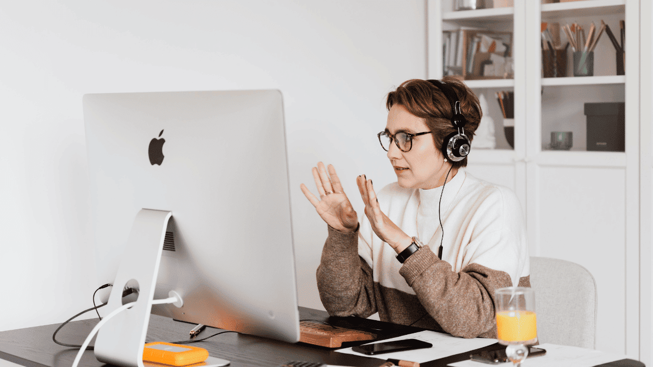 A person wearing glasses and headphones gesturing while sitting at a desk in front of a desktop computer trying to work.